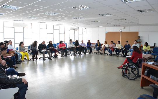 Foto de uma sala de aula com 24 pessoas (12 homens e 12 mulheres) sentadas em formato U, voltadas para a frente, onde há uma pessoa em cadeira de rodas. Ao fundo, uma parede formada com janelas de vidro refletindo a claridade do dia. Na outra parede, uma pessoa em pé e um quadro de cortiça. 